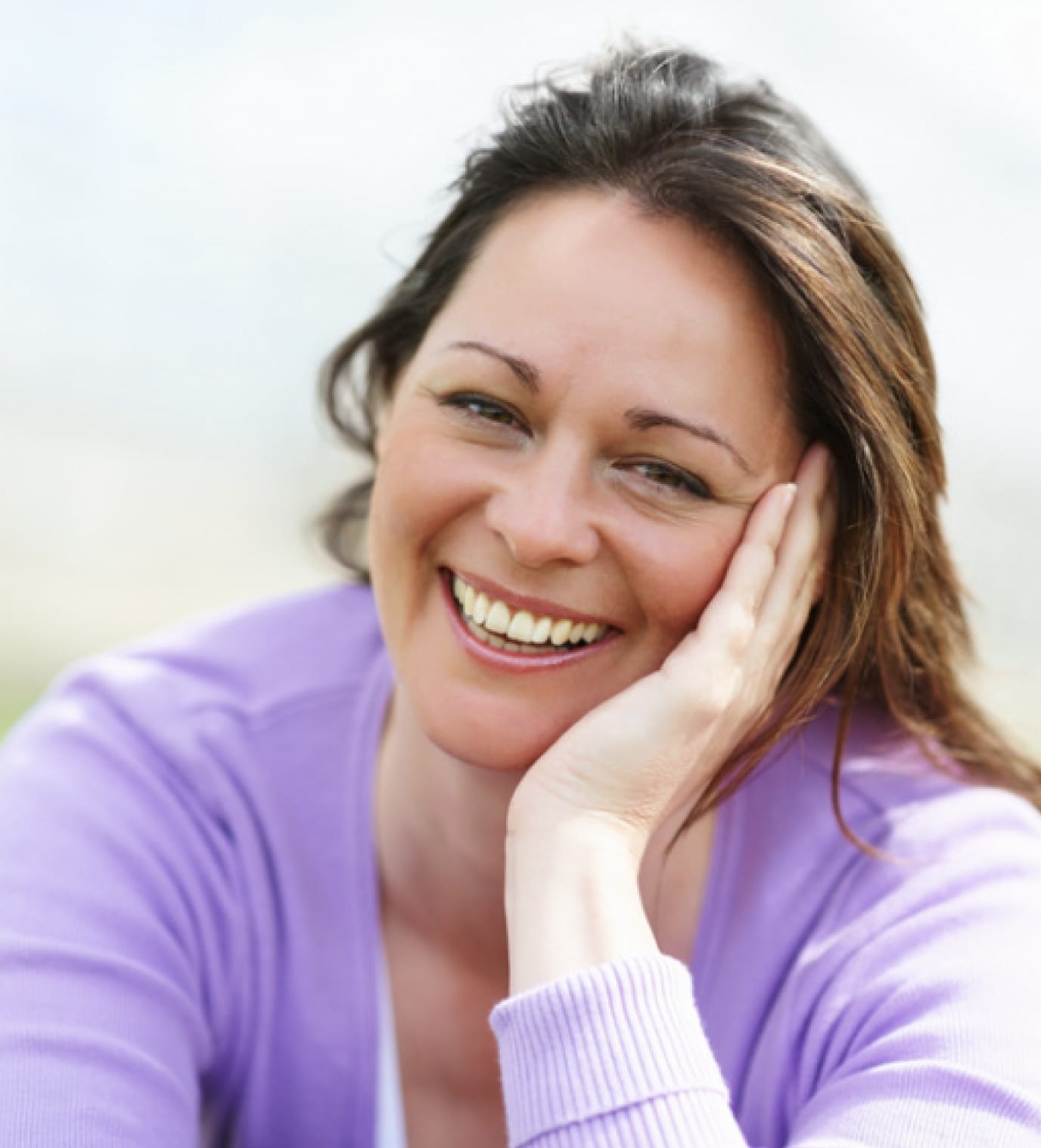 A woman with long brown hair smiles warmly at the camera, resting her face on her hand. She is wearing a light purple sweater and is outdoors with a blurred background.