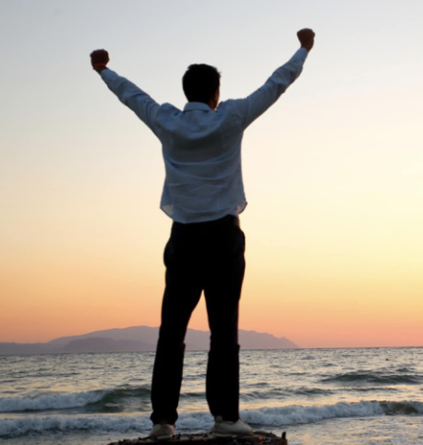 A person stands on the shore with arms raised in triumph, facing the ocean at sunset. The sky is orange and pink, and mountains are visible in the distance.