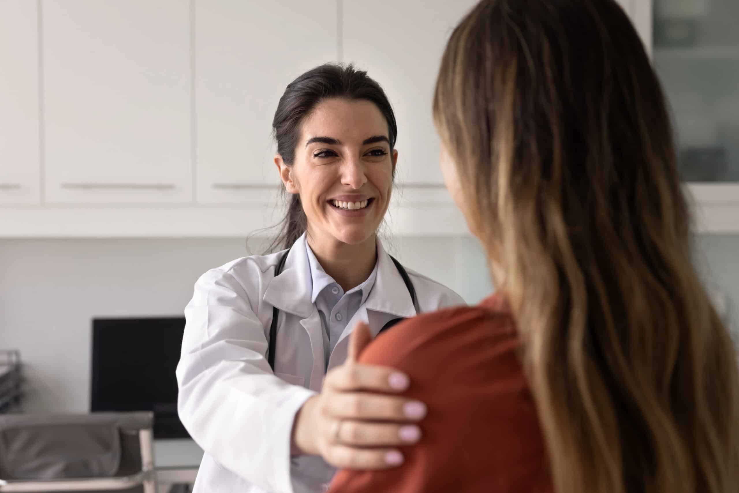 A smiling female doctor in a white coat reassures a patient by placing a hand on her shoulder in a medical office setting. The patient is seen from behind, with long hair and wearing a rust-colored top.