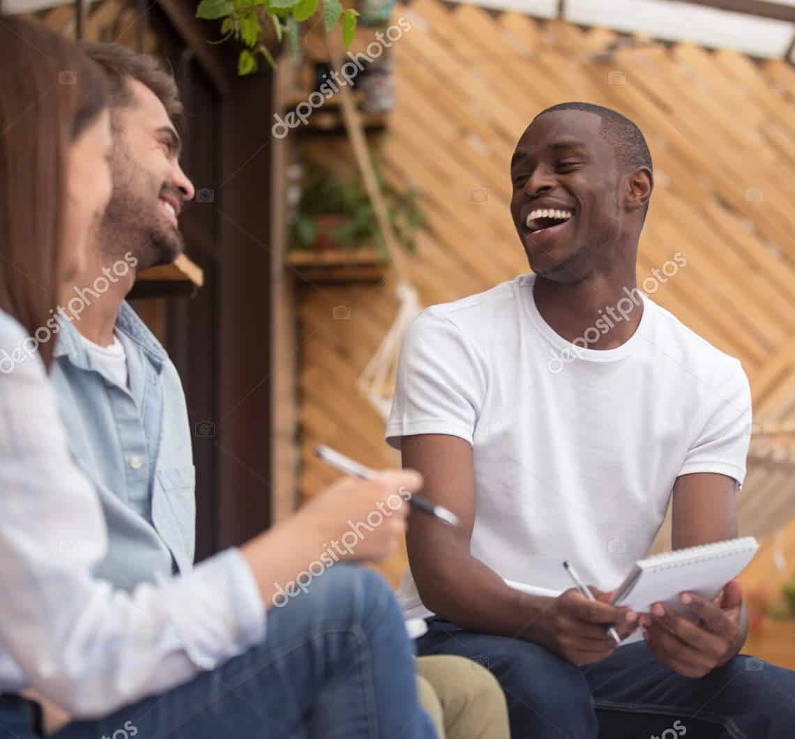Three young adults sit together outdoors, smiling and laughing as they engage in conversation. One person holds a notepad and pen, while others listen and enjoy the relaxed, friendly atmosphere.