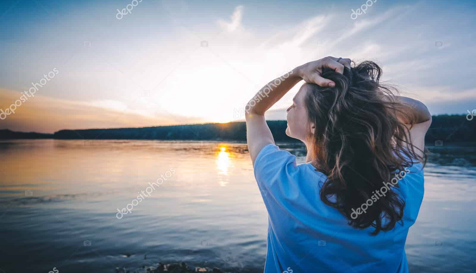 A woman with long brown hair stands by a calm river at sunset, holding her hair with one hand and looking toward the horizon, with trees visible in the distance under a colorful sky.