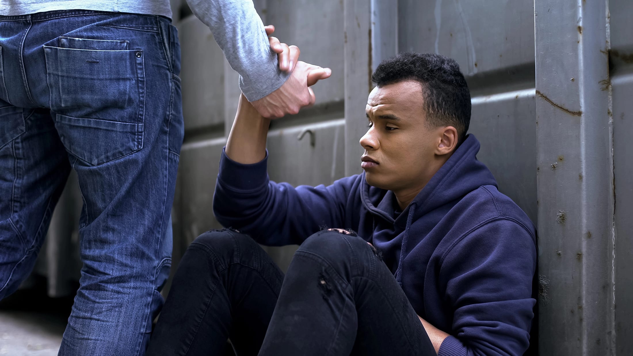 A person sitting on the ground against a wall is being helped up by another person offering a hand. The seated individual looks thoughtful and wears a navy hoodie and black jeans. The helper is standing and wearing blue jeans.