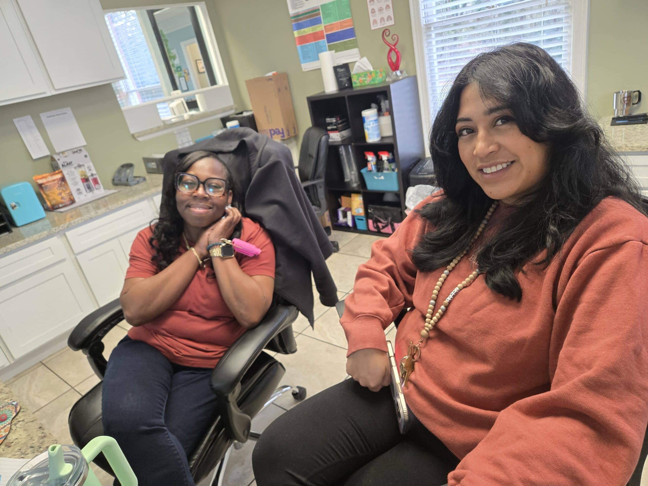 Two women sit smiling in an office, both wearing reddish tops. One is sitting back with her hands by her face, while the other leans forward holding a phone. Office supplies and shelves are visible in the background.