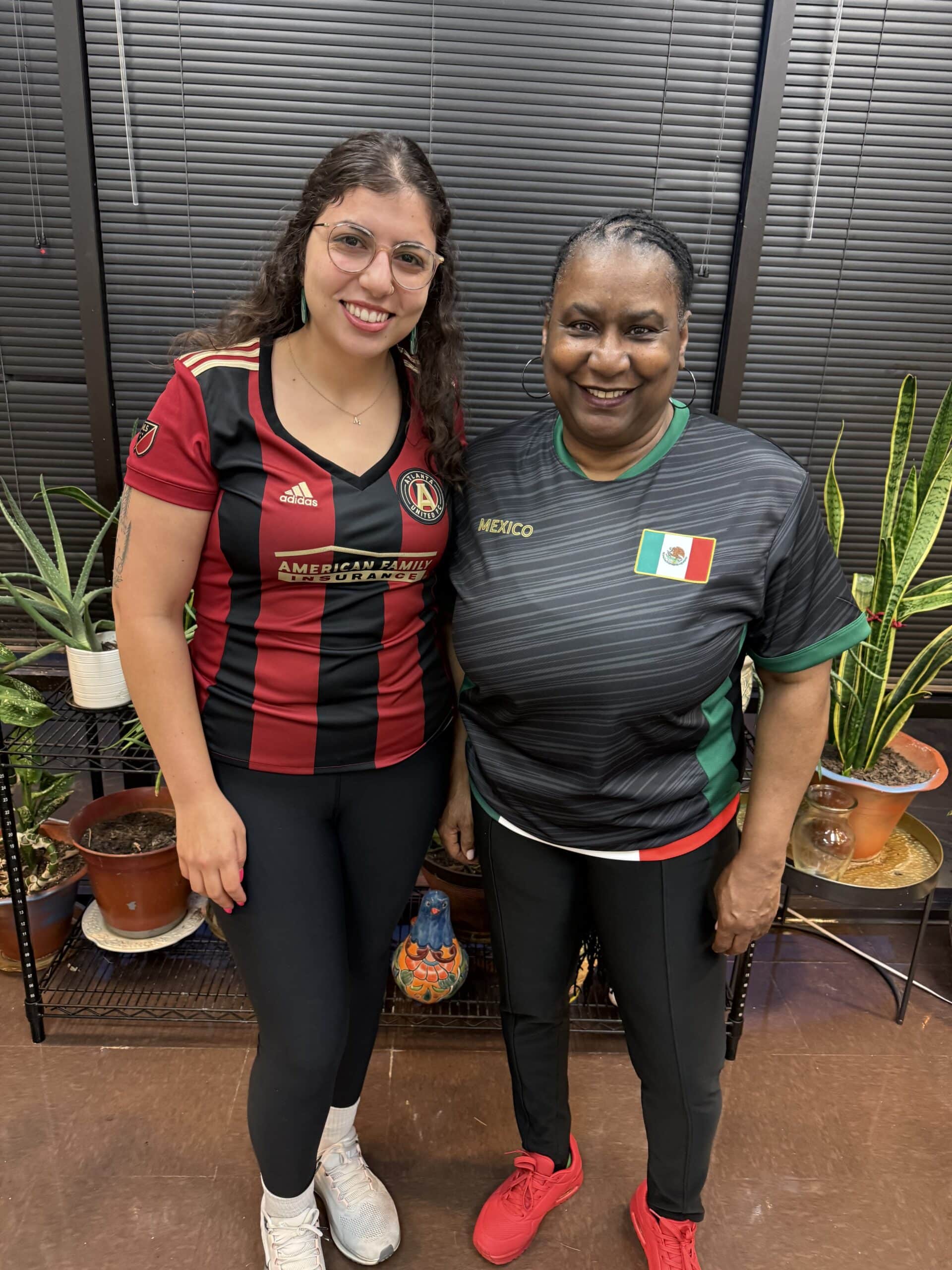 Two women smile and stand side by side indoors. Both wear soccer jerseys, one red and black, the other green with a Mexico flag. Several potted plants sit on shelves in the background.