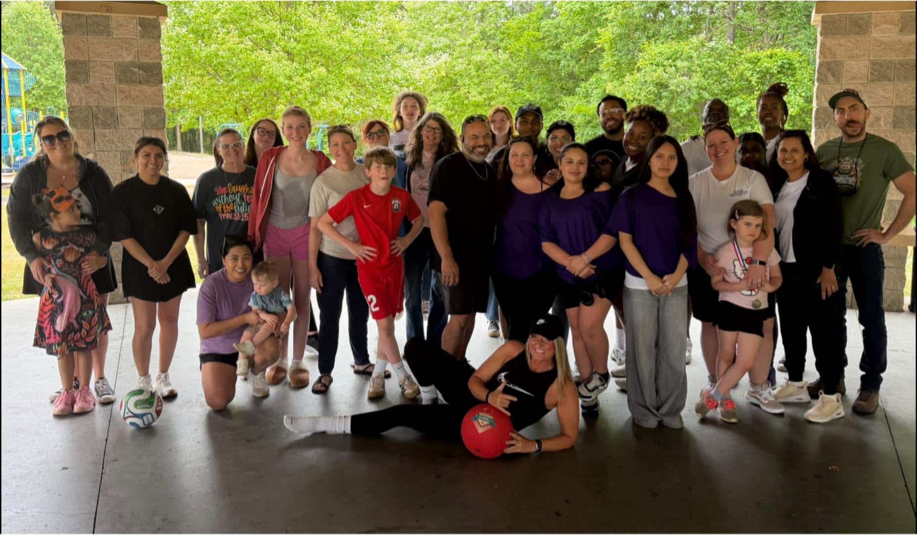 A diverse group of adults and children pose together under a pavilion, some wearing purple and sportswear. Trees and a playground are visible in the background. One person in front is leaning on a large red ball.