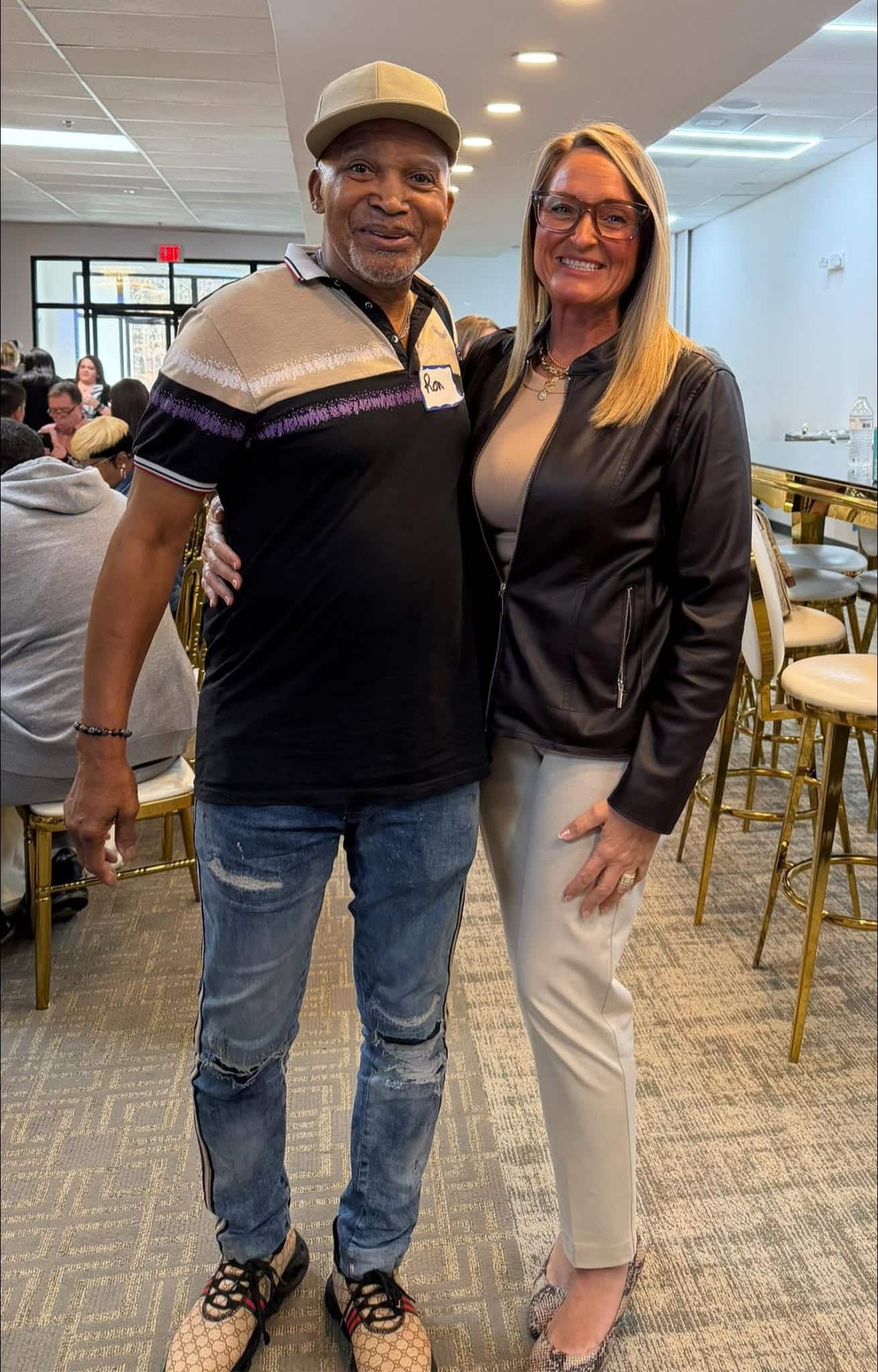 A man and a woman stand smiling together indoors at an event. The man wears a hat, black shirt, and jeans; the woman wears glasses, a black jacket, and light pants. People sit at tables in the background.