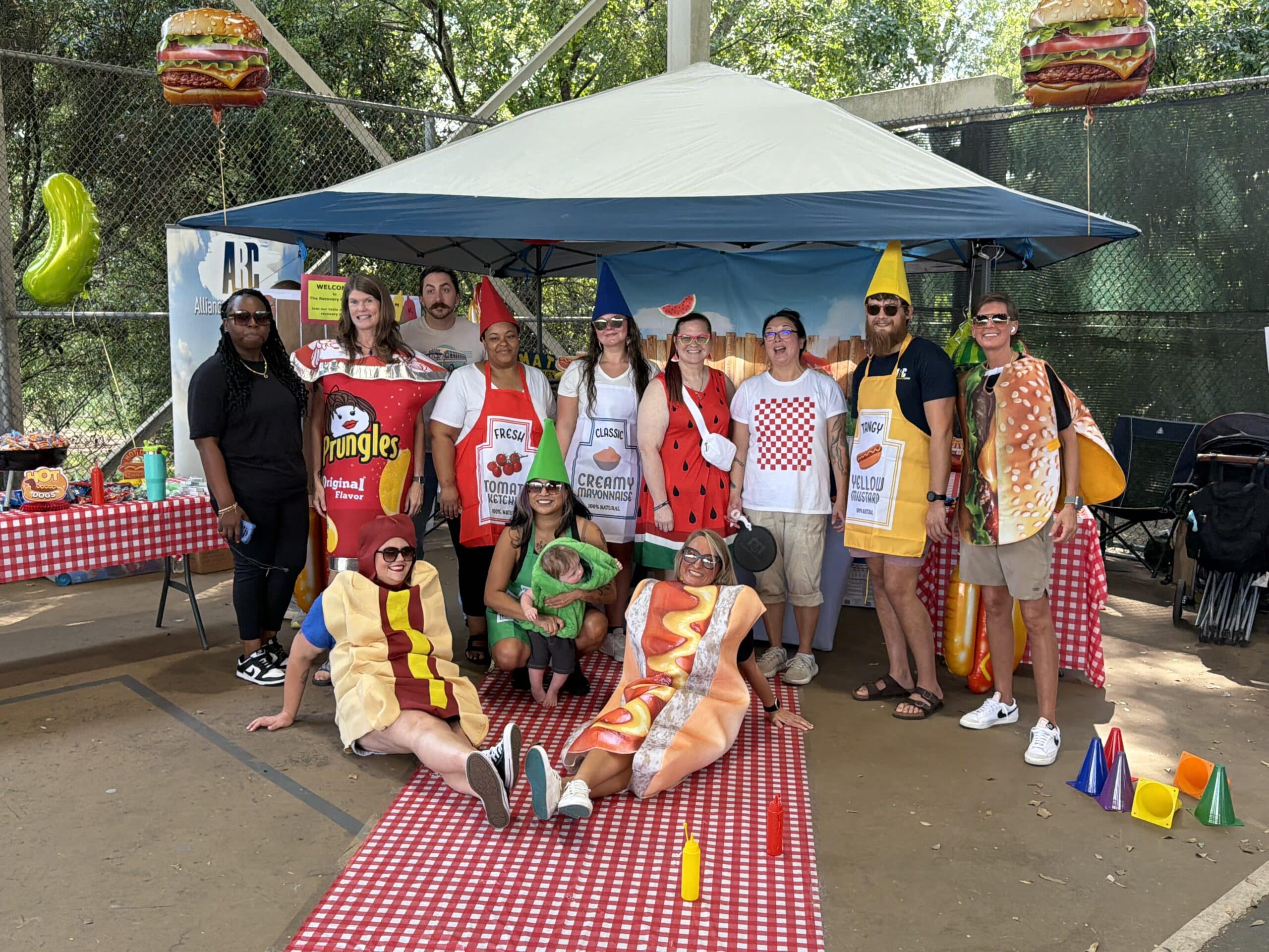 A group of people in colorful food costumes pose together in front of a tent with picnic decorations and a checkered tablecloth. Some are dressed as hot dogs, chips, ketchup, and mustard.