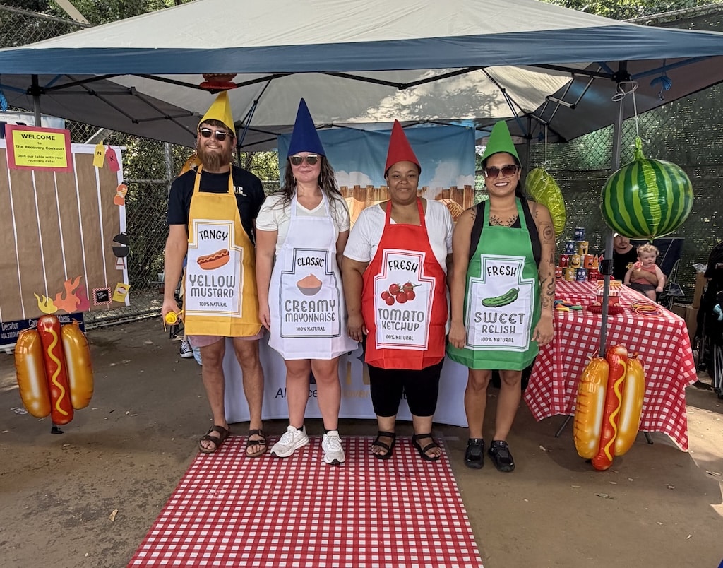 Four adults stand under a canopy, each wearing an apron and hat styled as mustard, mayonnaise, ketchup, and relish bottles. Picnic tables and hot dog decorations are visible around them.
