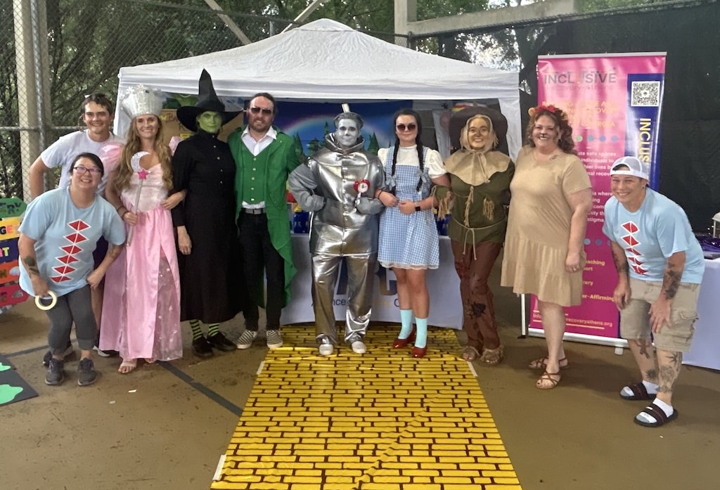 A group of people in Wizard of Oz costumes stand on a yellow brick road in front of a tent at an outdoor event, smiling at the camera.