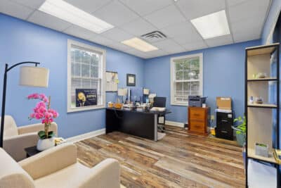A tidy office with blue walls, wooden floor, desk with computer, two beige chairs, pink flowers, bookshelves, windows with blinds, and a framed poster on the wall.