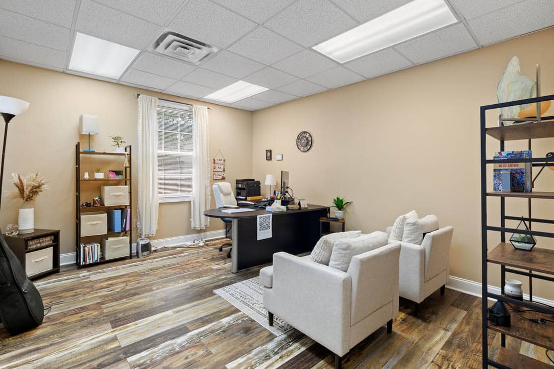 Modern office with beige walls, wood floor, a desk with computer, two white armchairs, shelves with decor, a large window with blinds, and ceiling lights.