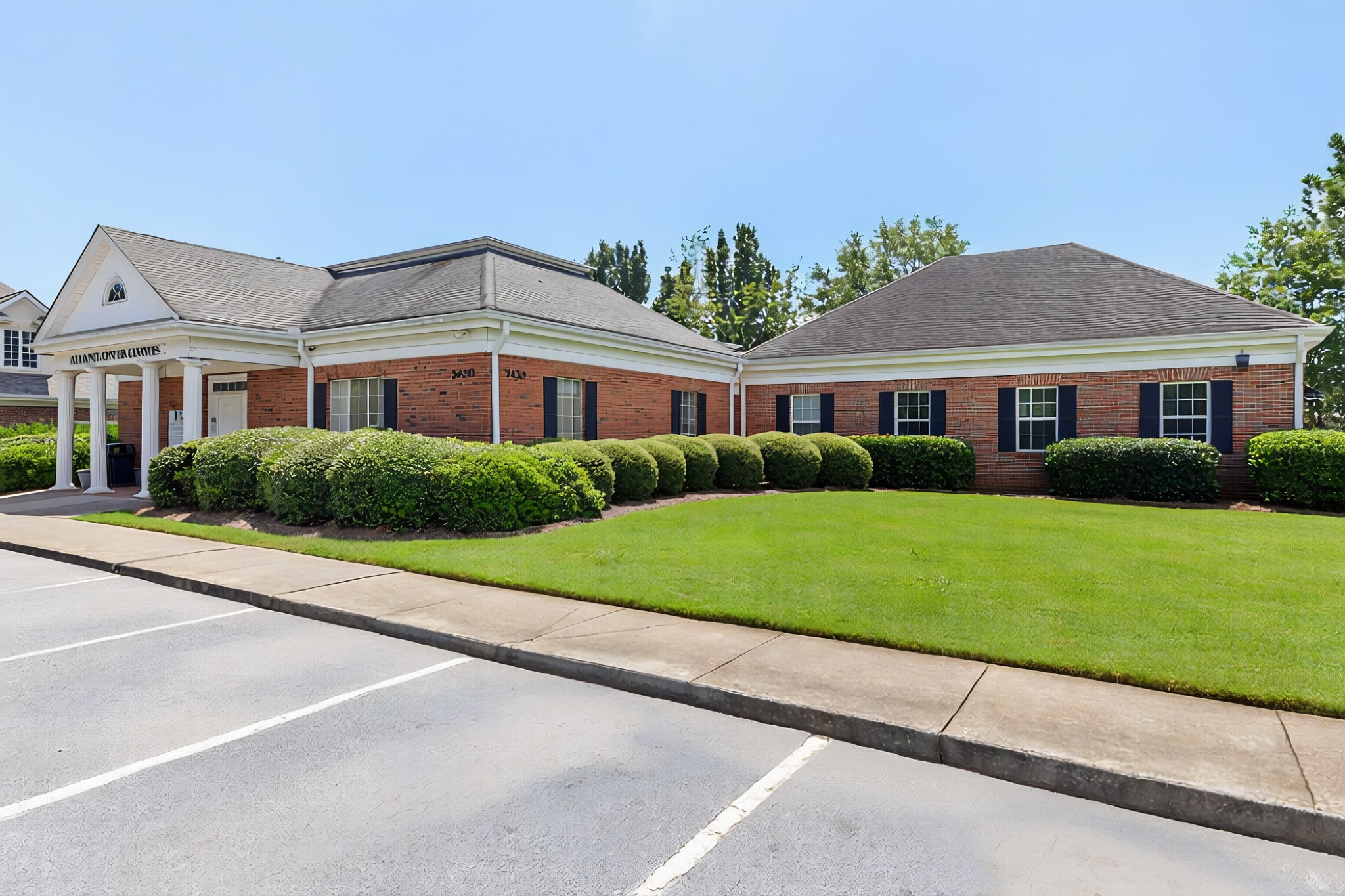 A one-story brick office building with white trim, surrounded by bushes and a neatly manicured lawn, sits next to an empty parking lot on a sunny day.