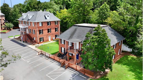 Aerial view of two brick buildings surrounded by trees, with a parking lot and empty parking spaces in front. One building is three stories, the other is two stories with a fenced walkway.