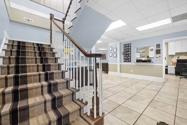 A tiled office lobby with a striped carpeted staircase, white railing, blue and white walls, bulletin boards, a reception window, and desks in the background under fluorescent lighting.