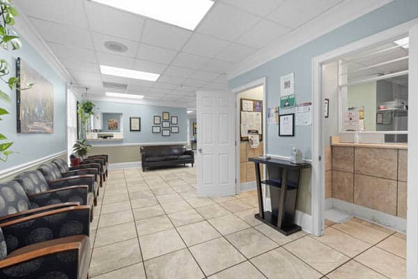 A clean, bright waiting room with patterned chairs along one wall, a black couch, tiled floor, wall art, and two open doors leading to office areas. A small table with sanitizer is near the doorway.