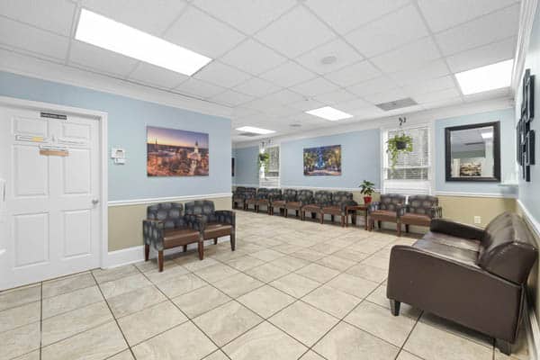 Spacious, well-lit waiting room with rows of chairs, a brown leather couch, light blue walls, tile flooring, and nature photos on the walls. Large windows let in natural light.