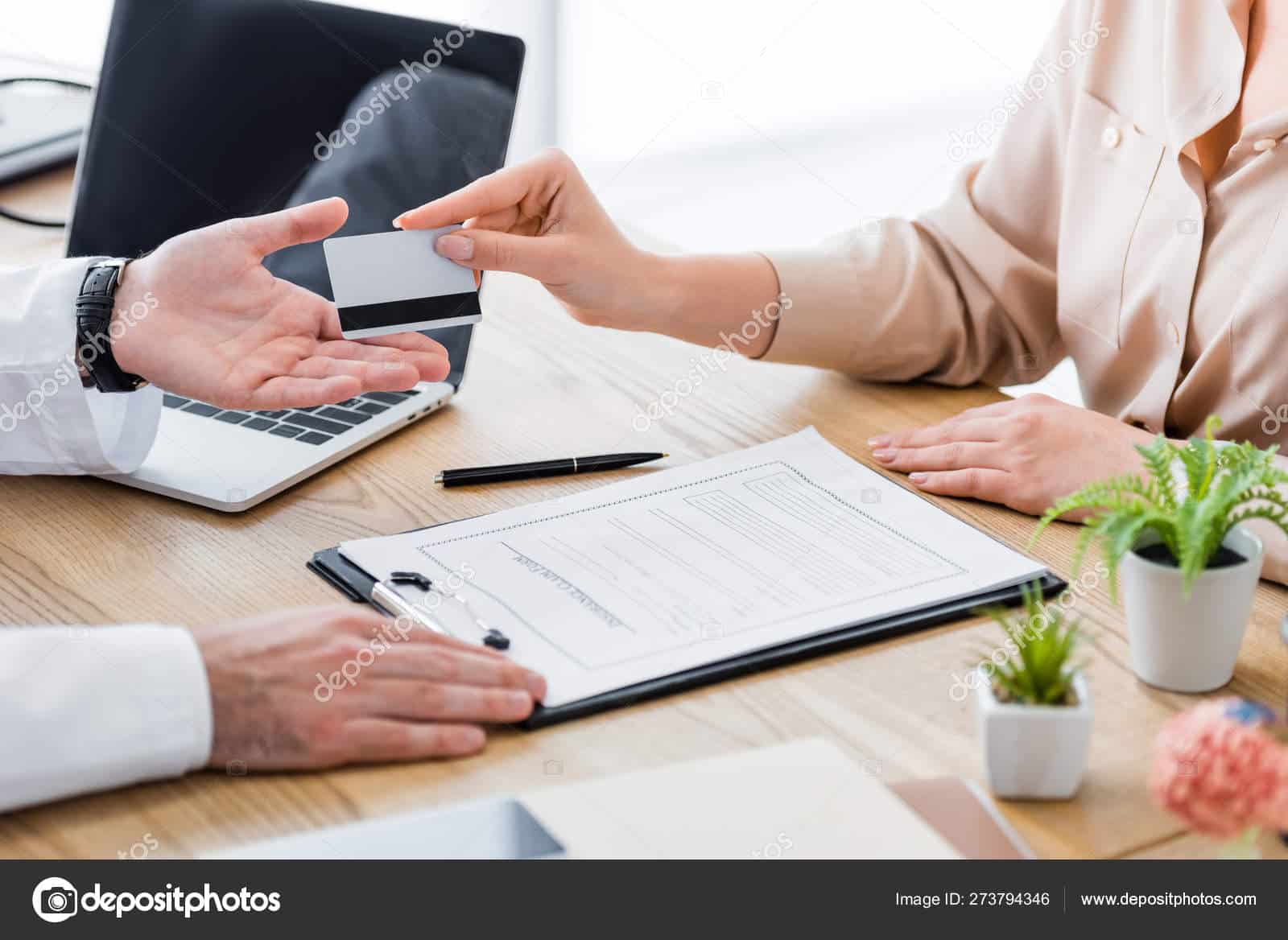 Two people sit at a desk; one hands a card to the other. A clipboard with a document, pen, laptop, and small potted plants are visible on the wooden surface. Only their hands and part of their arms are shown.