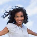 A woman with curly hair smiles outdoors against a blue sky with clouds. She wears a light-colored top and appears happy and carefree.
